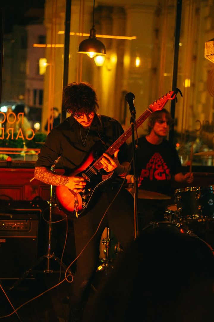 Jack playing guitar and singing in front of a big window with the street lights behind him, a drummer to his side